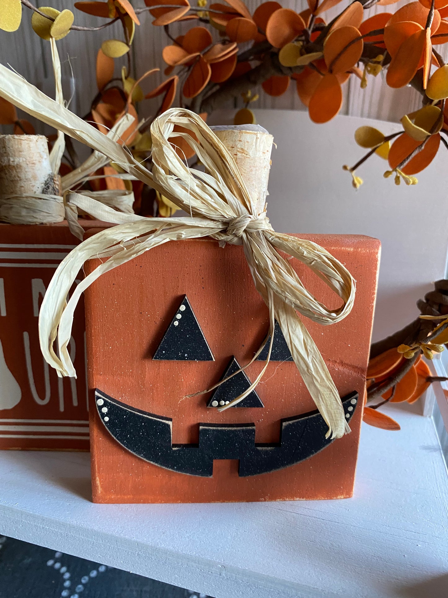 A reversible pumpkin-shaped sign with a jack-o-lantern face on one side and a text 'Oh My Gourd' on the other, decorated with a beige ribbon, placed on a table with autumnal surroundings.