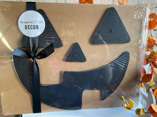 A packaged No Carve Jack-o-lantern Kit with cut-out shapes for crafting a pumpkin decoration, Tooth Mouth style, tied with a black ribbon, against a fall-themed backdrop.