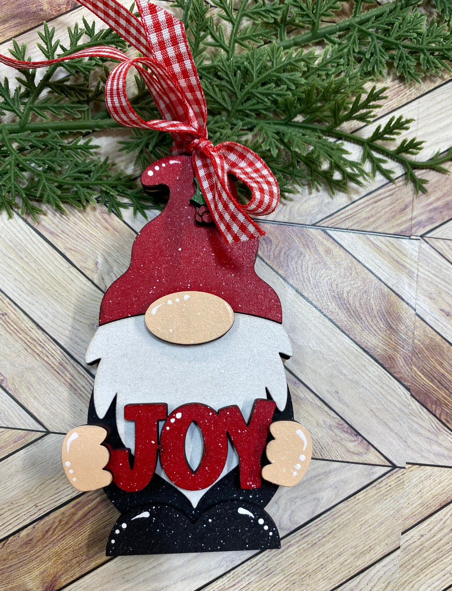 A gnome ornament with a red hat and black and white outfit, holding a sign with the word 'JOY' in front of a Christmas tree.