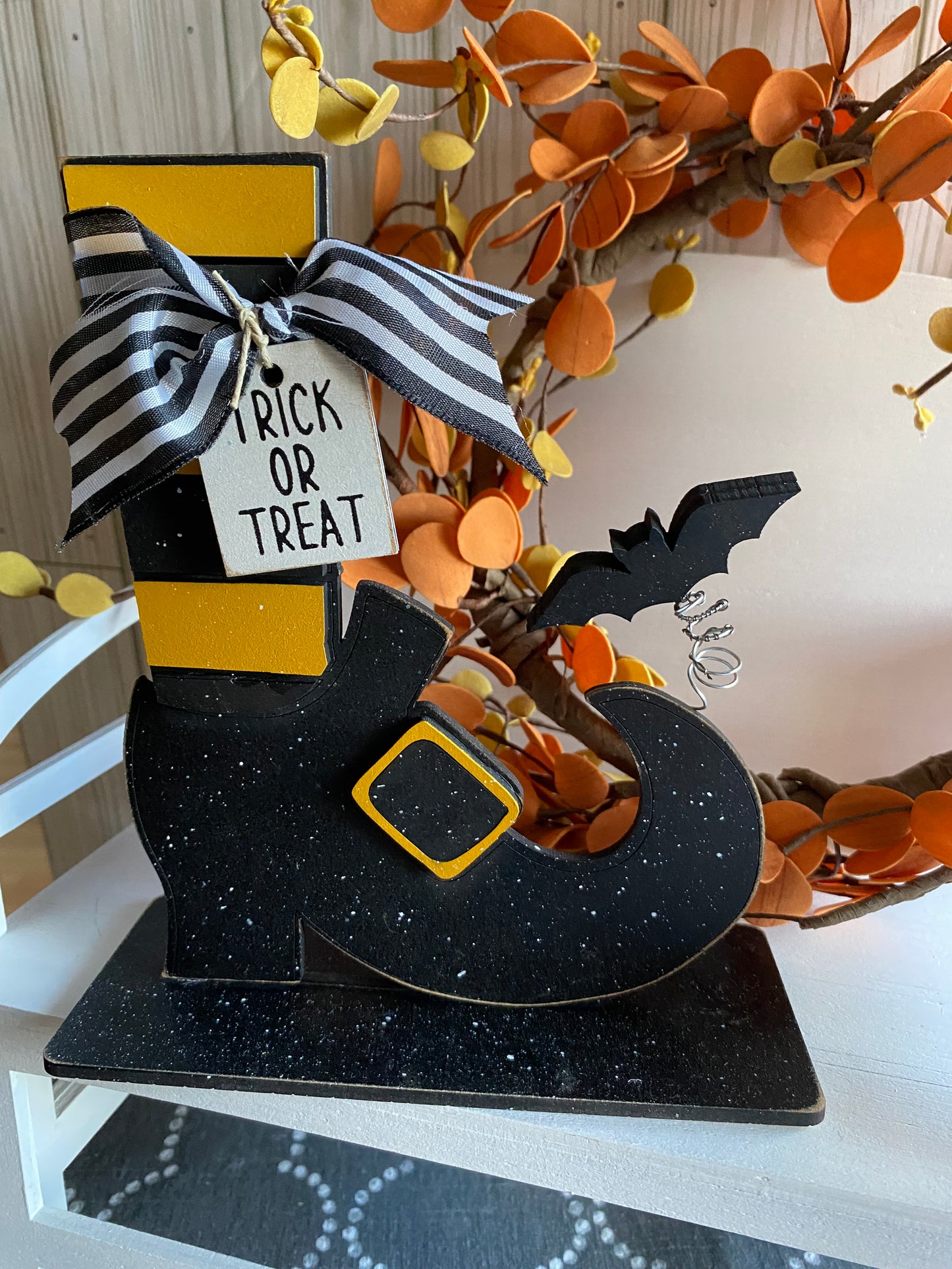 A decorative witch shoe with a 3D bat attached to it, painted in black and gold with a 'Trick or Treat' tag, displayed against a seasonal decor background.