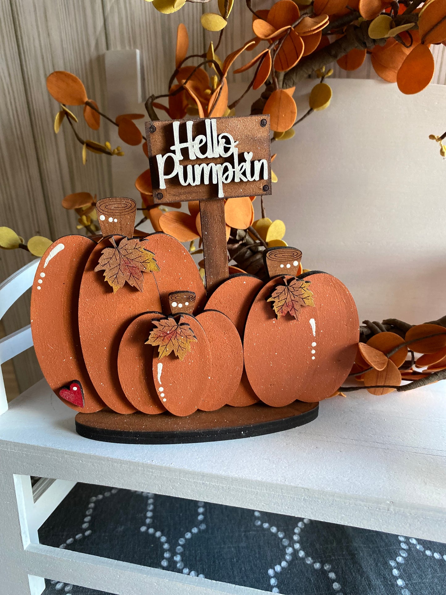 A hand painted decorative pumpkin with the text 'Hello Pumpkin' written on top, arranged with other pumpkin shapes and leaves in the background, indicating a fall theme.