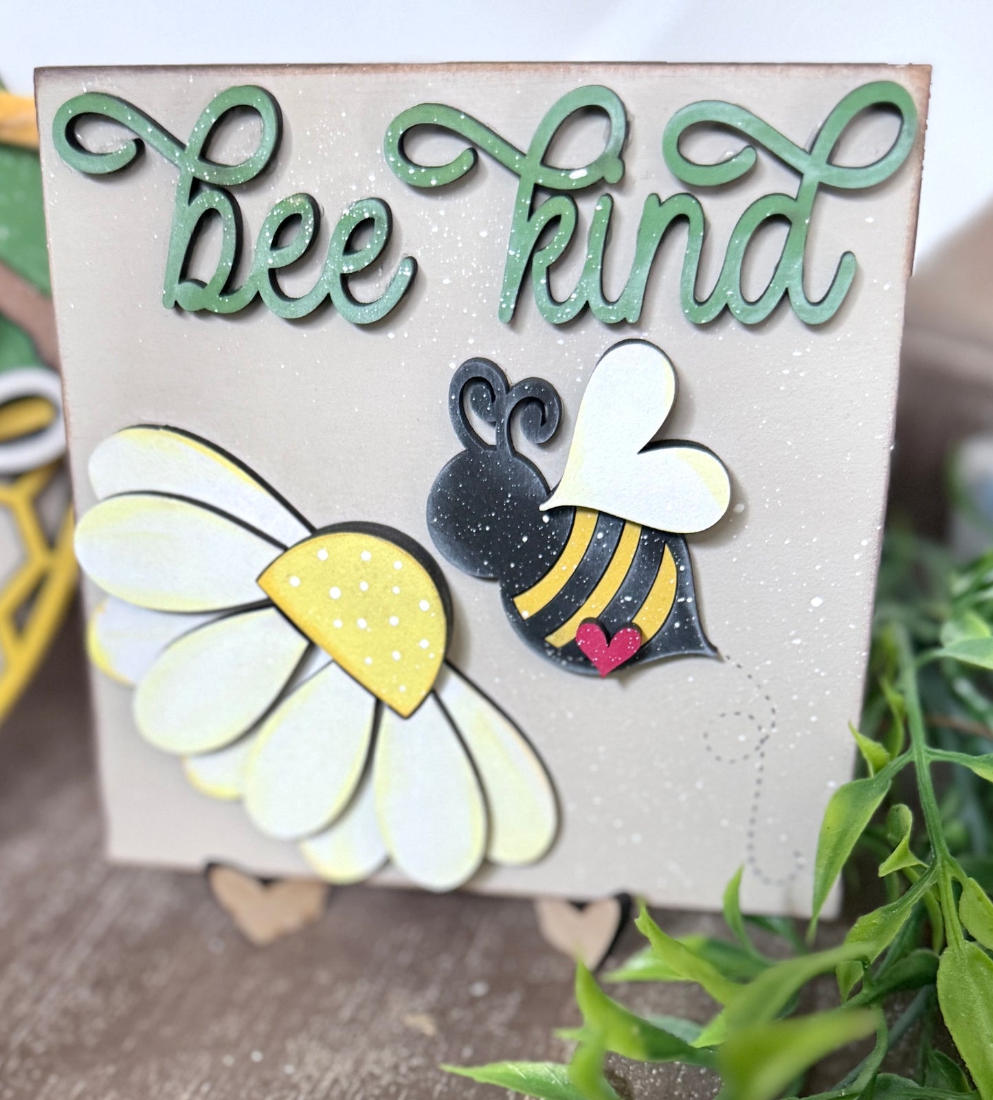 A set of three decorative signs with bee-themed designs and the phrase 'Bee Kind' on them, displayed on a tiered tray with a green plant in the background.
