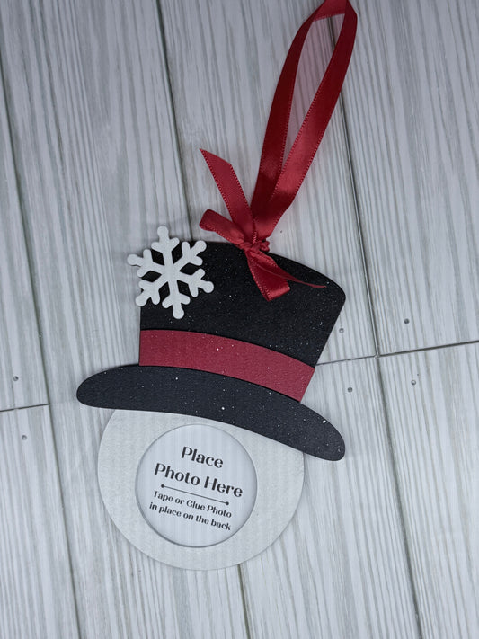 Decorative photo frame shaped like a snowman with a black top hat, red ribbon, and white snowflake on a light wooden surface.