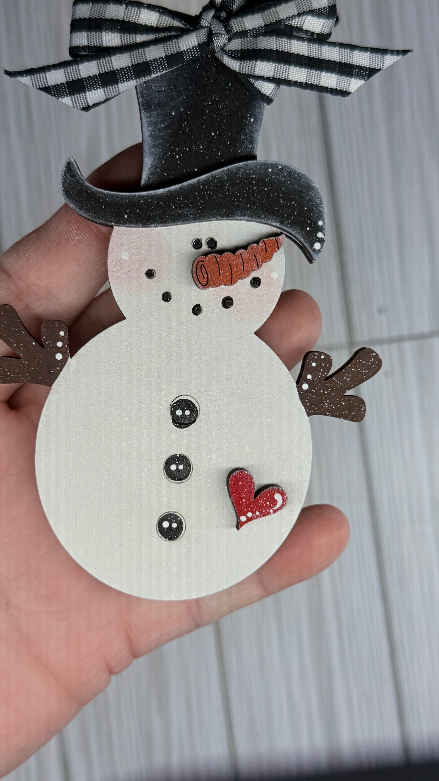 Hand holding a small snowman ornament with a black top hat and red heart on a light wooden background.