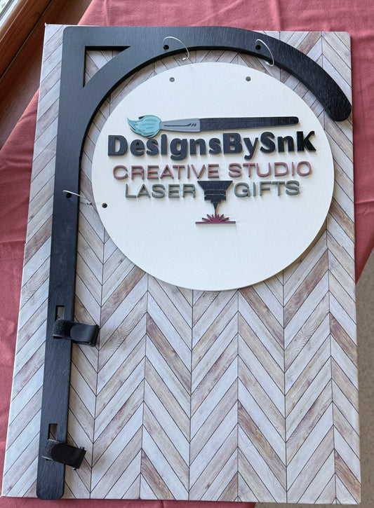 Wooden sign with 'DesignsBySnK' branding in 3D on a black tent bracket.  