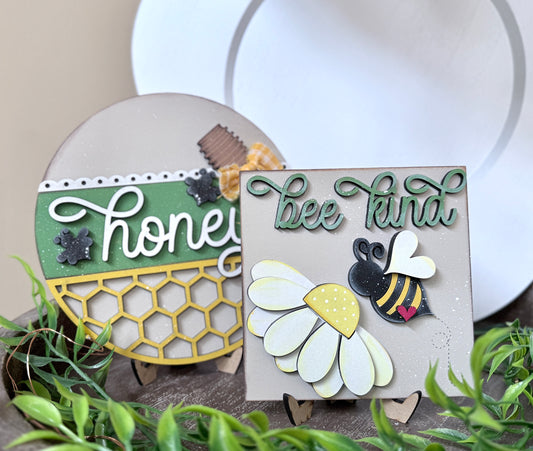 A set of two decorative signs with bee-themed designs and the phrase 'Bee Kind' & "Honey" on them, displayed on a tiered tray with a green plant in the background. Also included is a matching mini banner and easel  