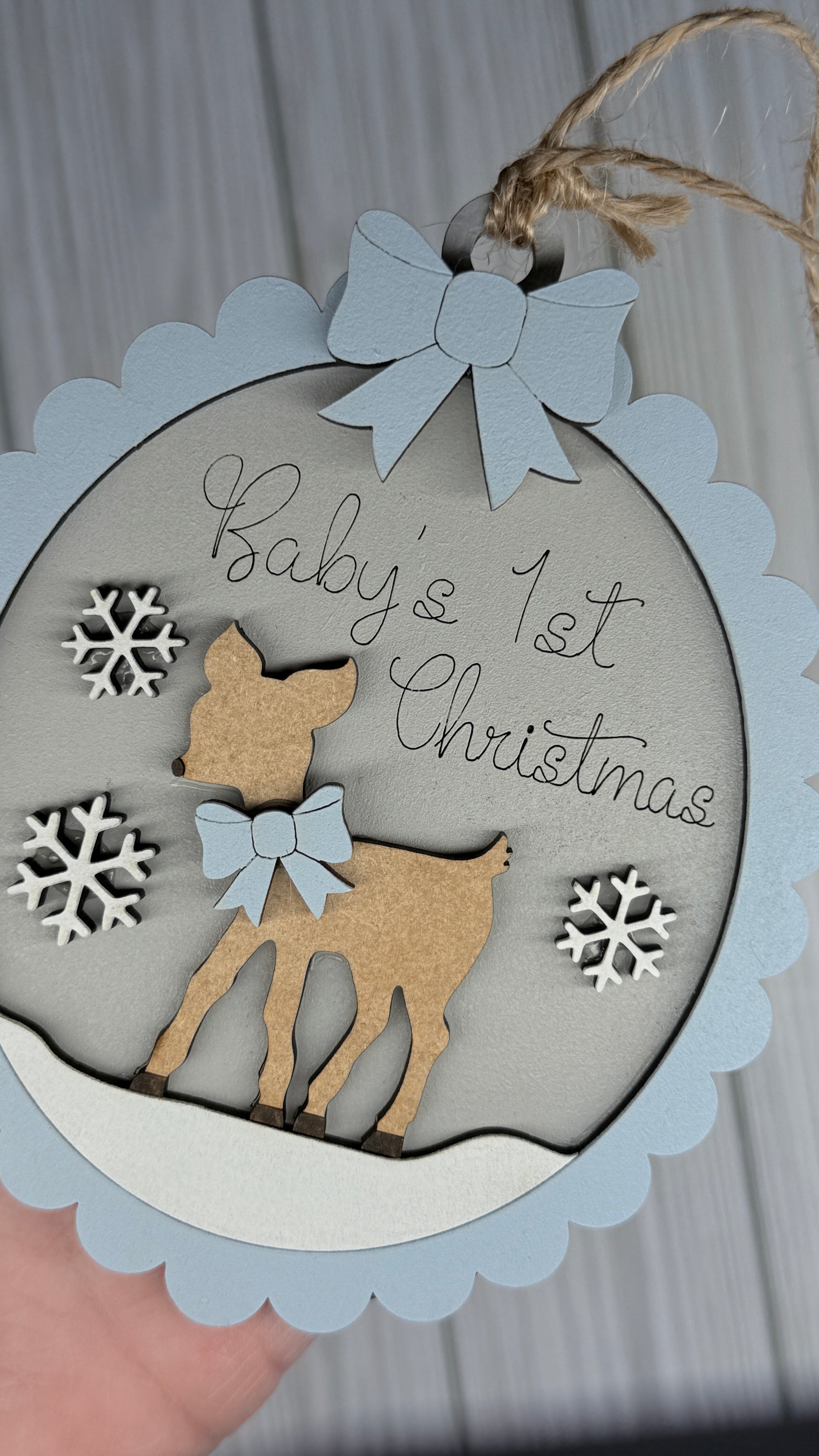 Decorative ornament with deer and snowflakes for 'Baby's 1st Christmas' on a light gray background. and framed in blue
