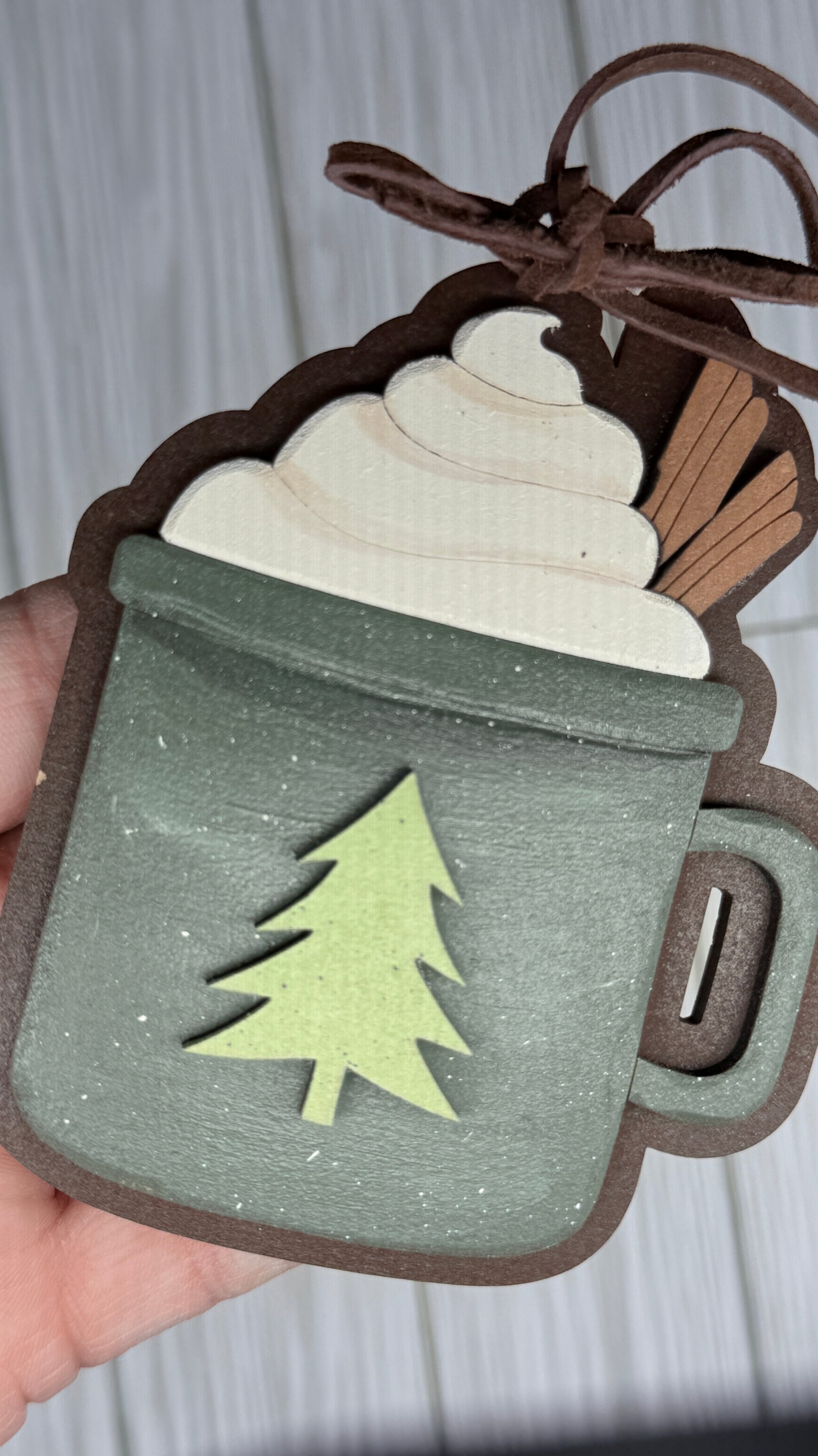 Decorative mug ornament with a tree cutout held by a hand against a neutral background. Close up for details and Leather bow and hanger. 