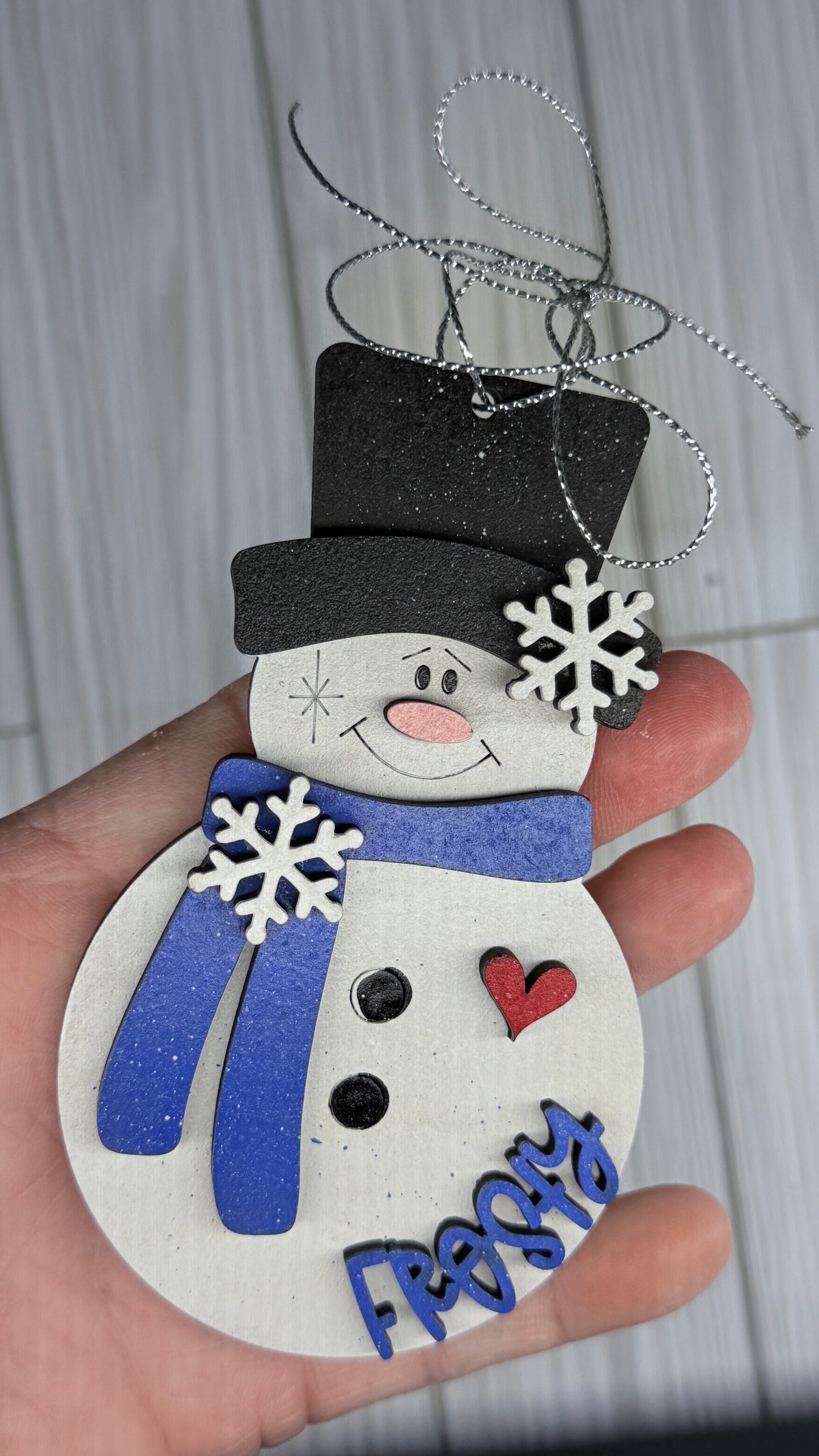 Hand holding a snowman ornament with a blue scarf and black top hat against a light background