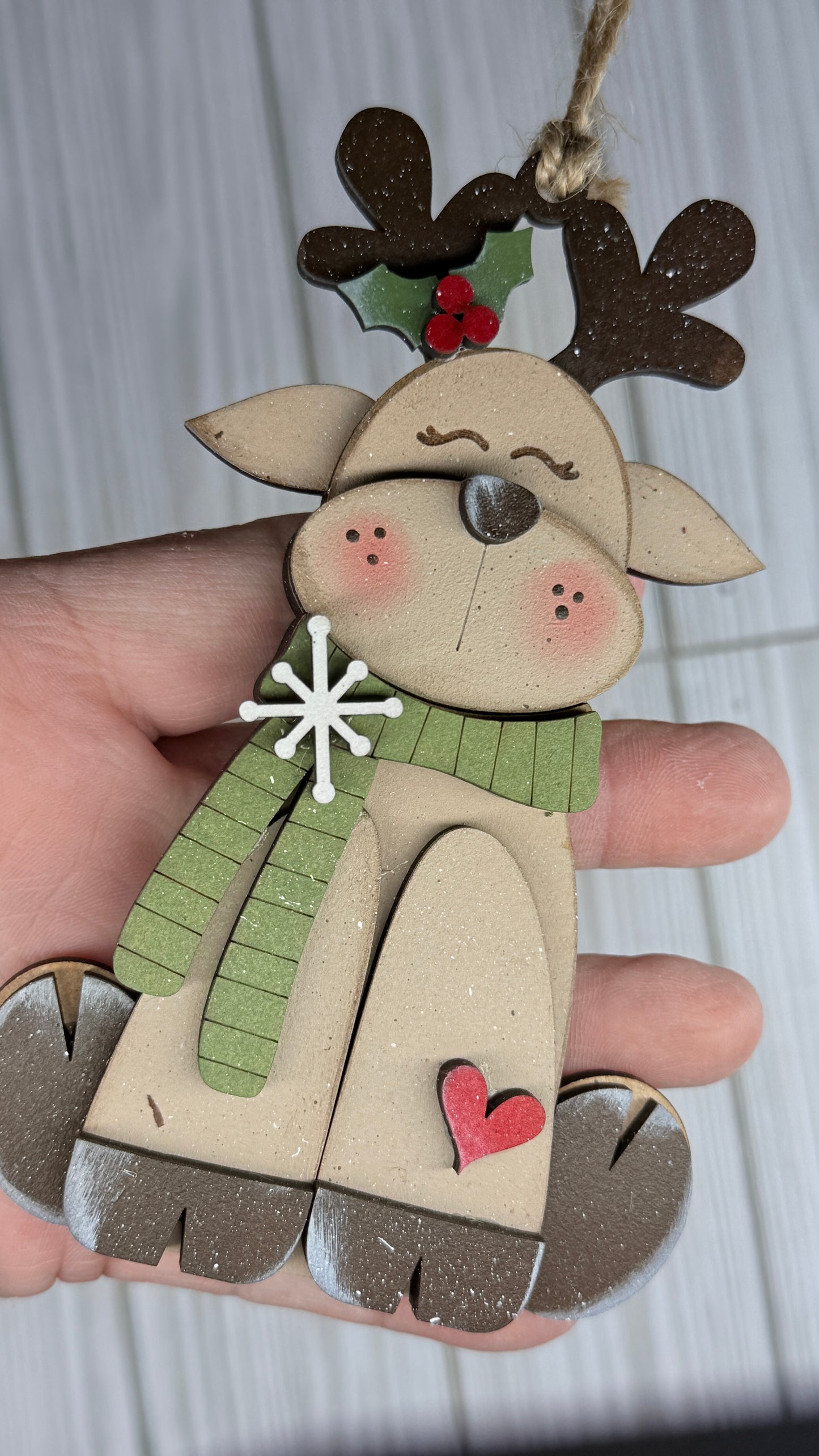 Wooden reindeer ornament with a green scarf, red heart, and holly leaves held by a hand against a light background.