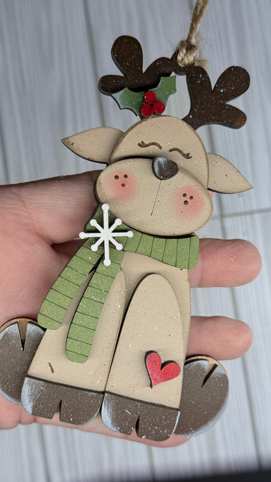 Wooden reindeer ornament with a green scarf, red heart, and holly leaves held by a hand against a light background.