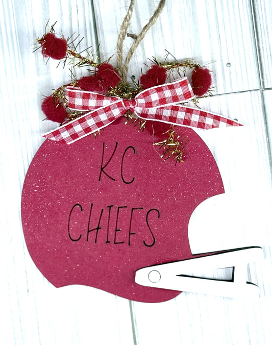 Decorative ornament shaped like a football helmet with 'KC Chiefs' text, red pom-poms, and a checkered ribbon.
