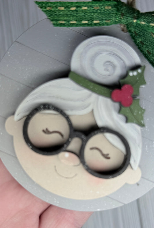 Grey shiplap round, Mrs Clause with a holly leaf in her hair, wearing black frame glasses and a rosy nose. 