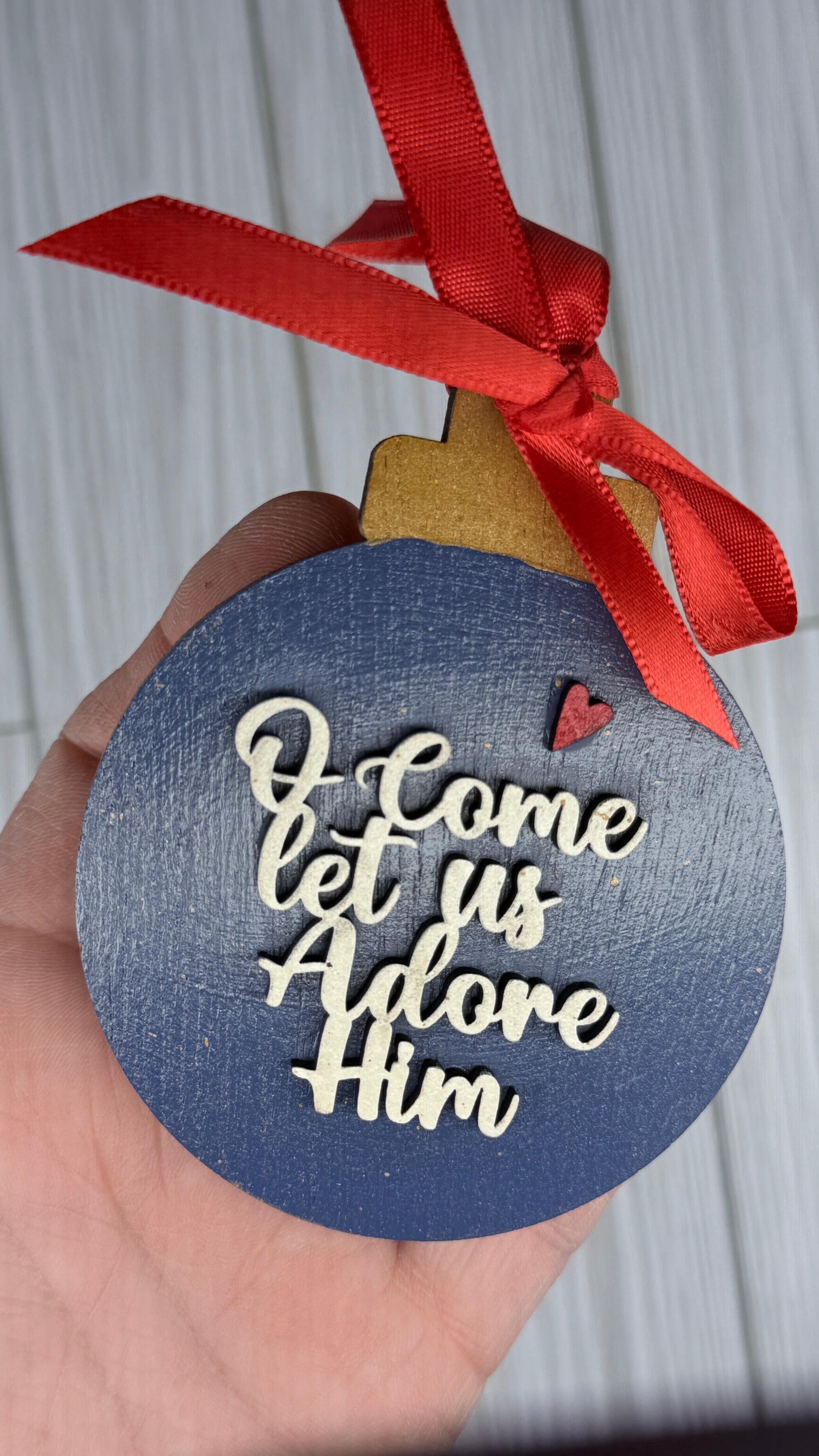 Decorative ornament with 'O Come Let Us Adore Him' text and red ribbon.