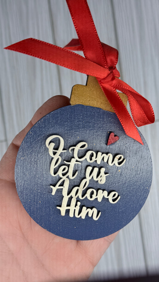 Decorative ornament with 'O Come Let Us Adore Him' text and red ribbon.