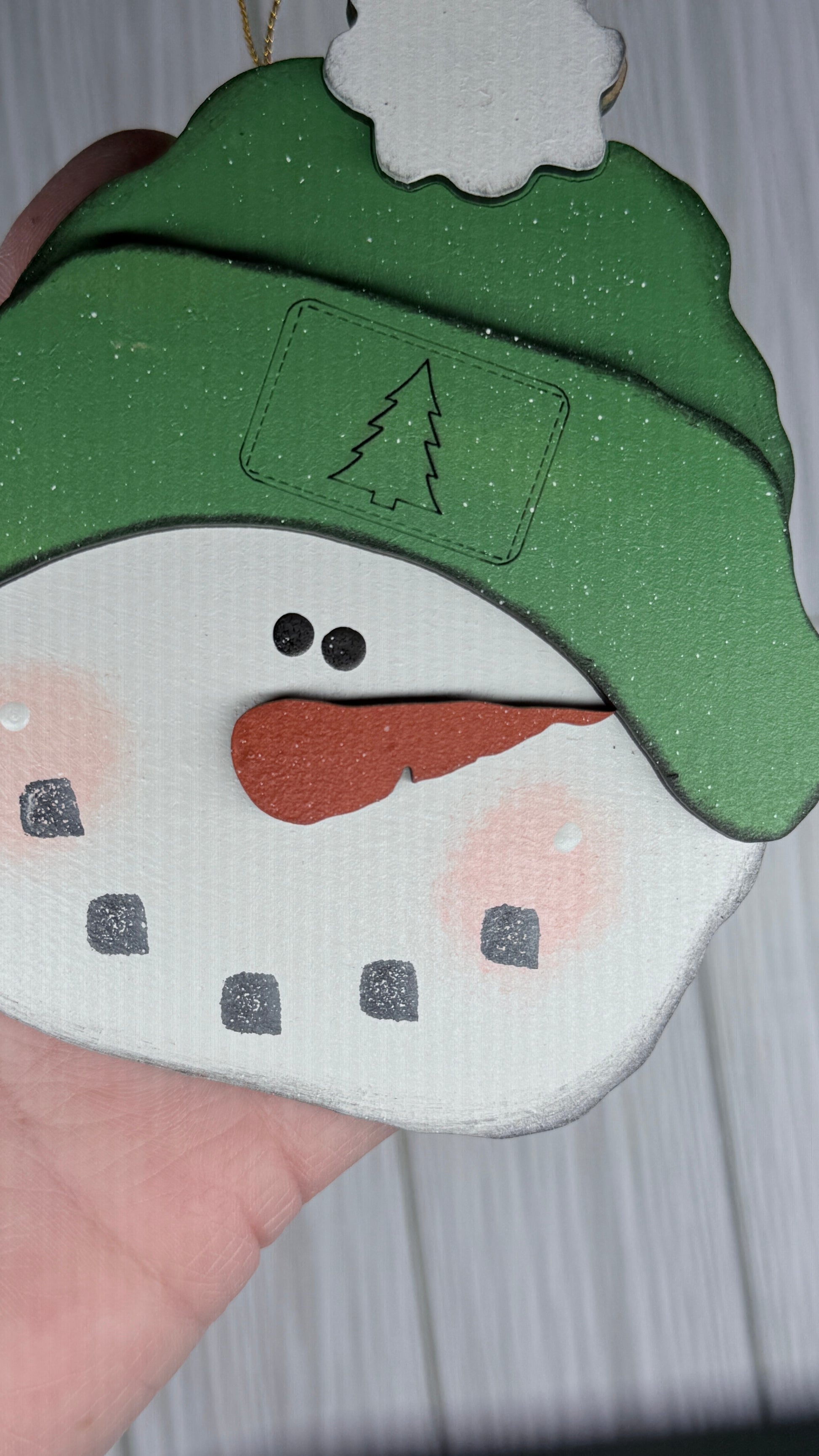 Snowman-shaped ornament with green hat and orange carrot nose held by a hand for close up detail workmanship.