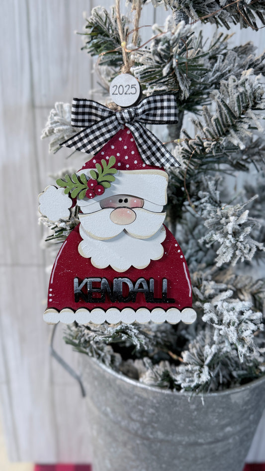 Decorative Santa Claus ornament with a checkered bow on a Christmas tree. Kendall on front.