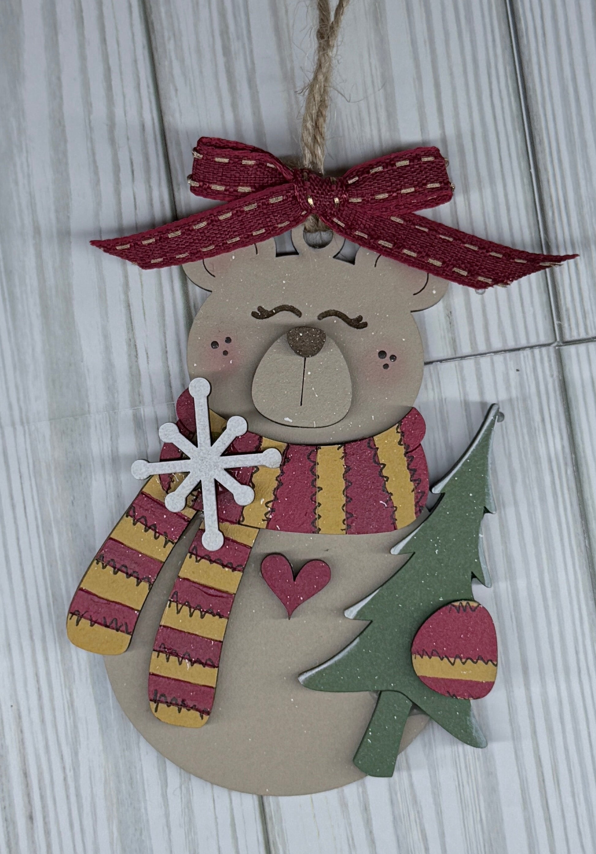 Decorative hanging ornament with a bear design, striped scarf, and red and gold bow on a wooden surface.