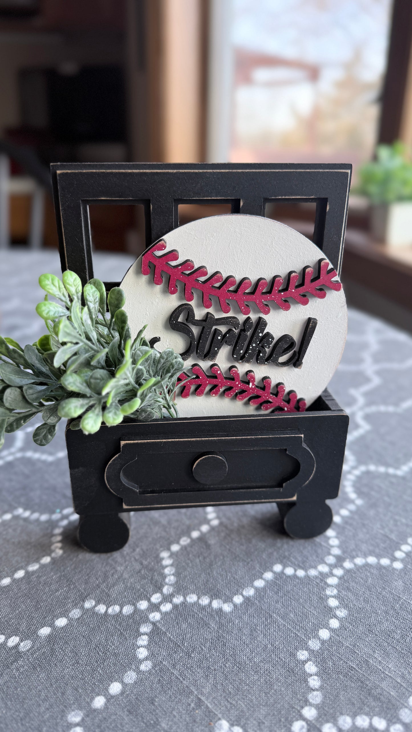 Decorative item with 'Striked' on a baseball design, placed on a black stand with greenery.