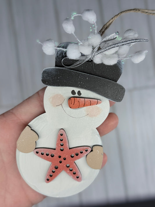 Decorative snowman ornament with a starfish and a black hat held by a hand against a neutral background.